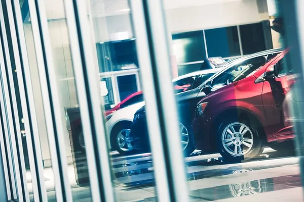 A view through glass panels showcasing a red car and several other vehicles inside a modern car dealership.