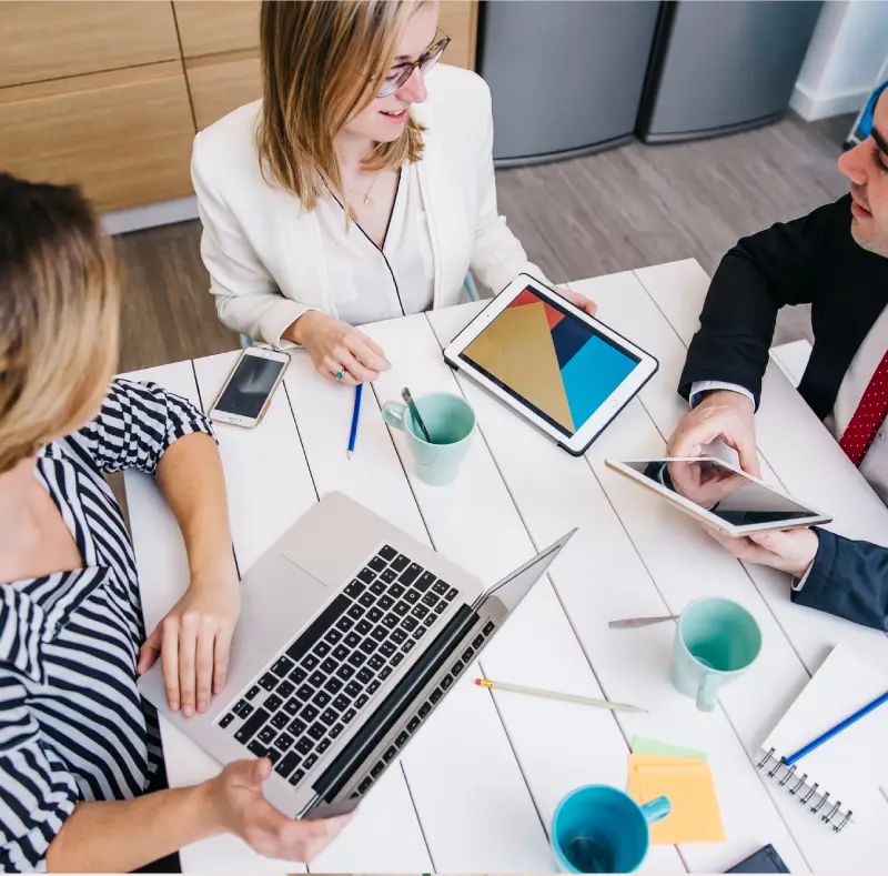 Three professionals engage in a digital discussion at a white table, surrounded by laptops, tablets, phones, and notepads.